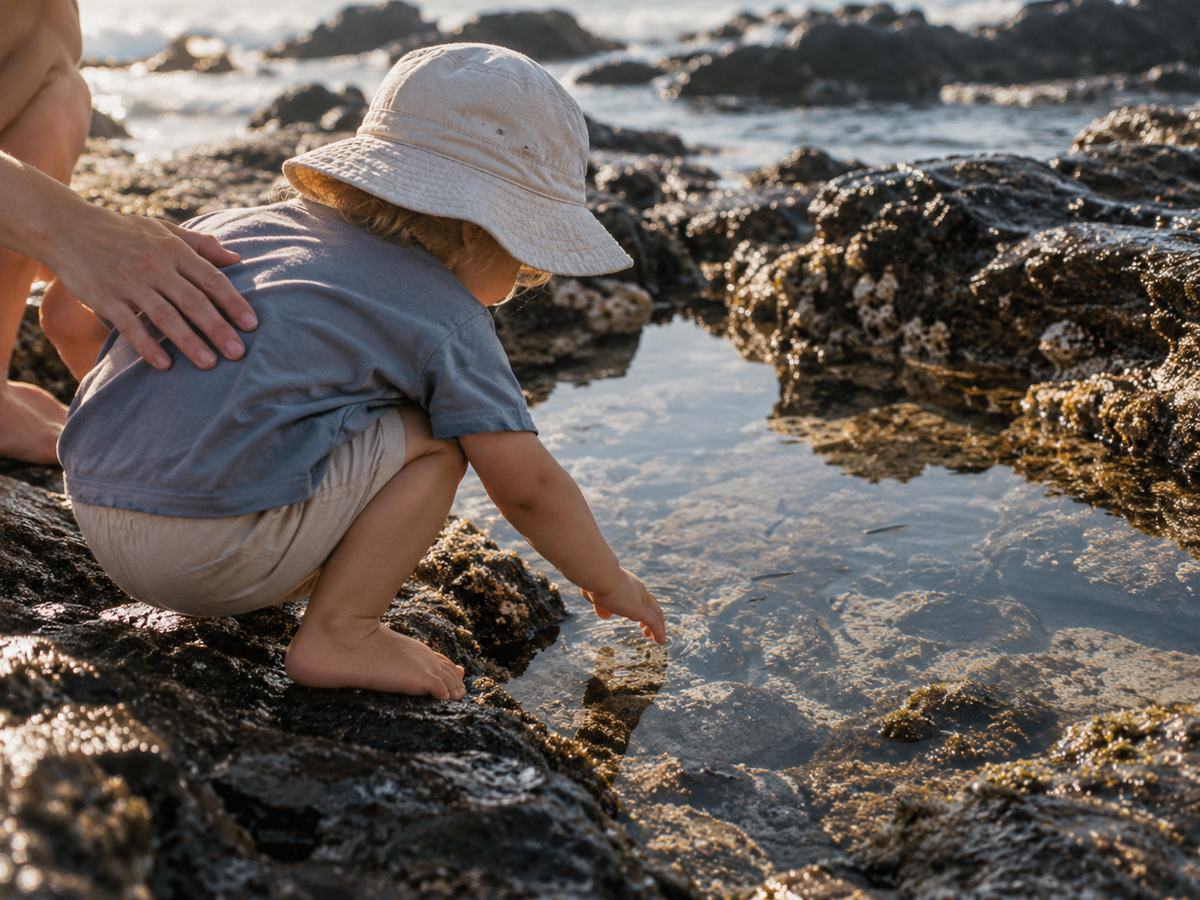 A toddler in a canvas hat crouches by a warm rocky pool, a parent's hand supporting their back.